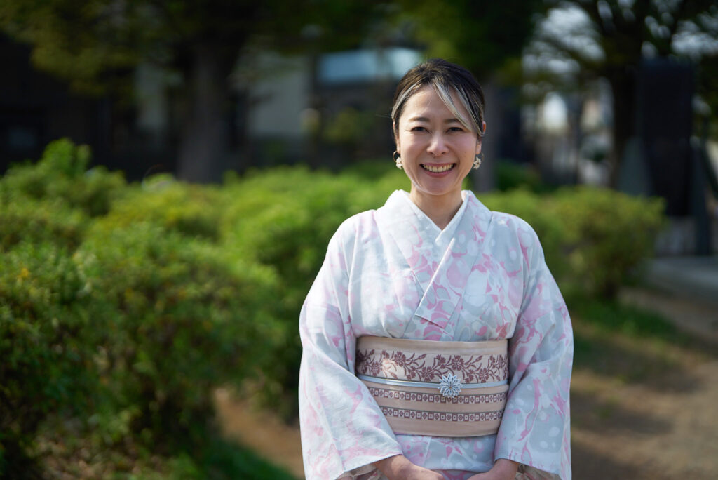 A smiling woman in a pale pink floral summer kimono standing in a sunlit park, the tsumami-zaiku artisan featured in the interview