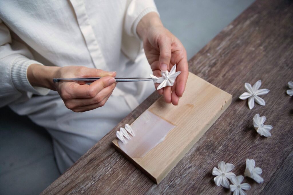 Close-up of hands using metal tweezers to fold a small white silk petal into a tsumami-zaiku flower on a wooden board, with finished cloth blossoms nearby