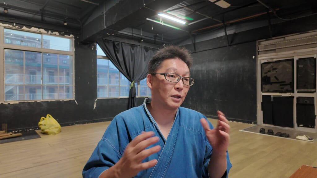 A Japanese man with glasses in a blue training jacket gesturing with both hands as he speaks in a dojo with large windows