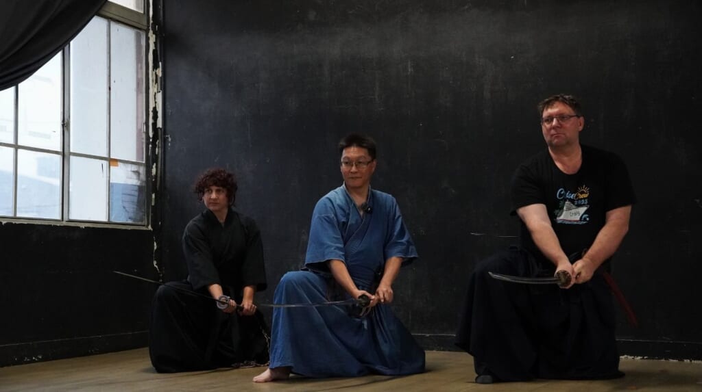 A Japanese instructor in a blue training uniform kneels on one knee holding a katana, flanked by two students also holding swords