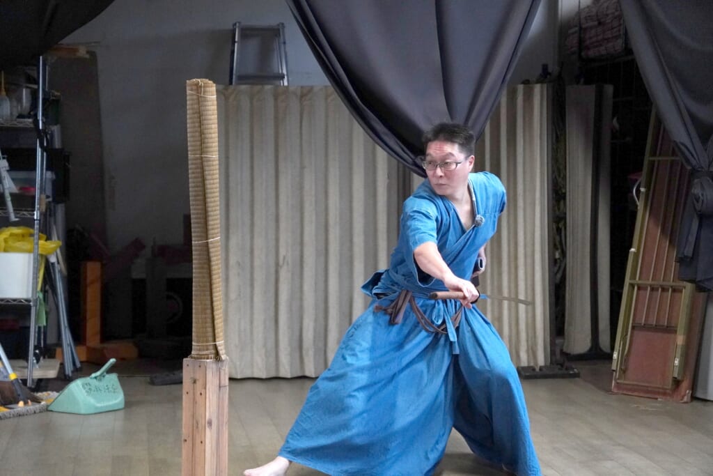 Gensei Kamiide in a blue training uniform in a low stance with his sword drawn beside a rolled tatami target in a dojo