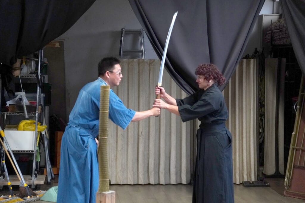 Gensei Kamiide in a blue training uniform steadies a student's grip on a raised katana during a battodo lesson