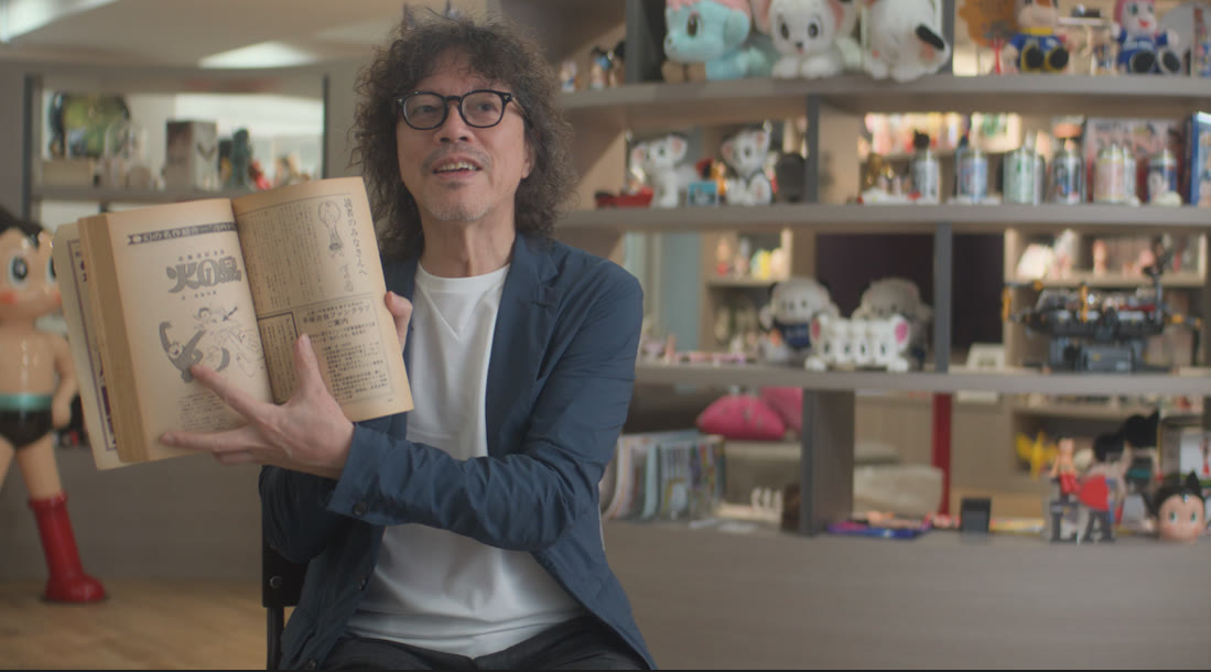 A man with curly hair and glasses holding open an old manga volume in a studio lined with toys and figures