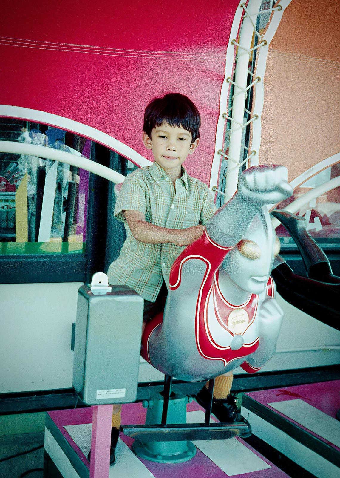 A young boy in a green checked shirt sitting on a red and silver Ultraman kiddie ride