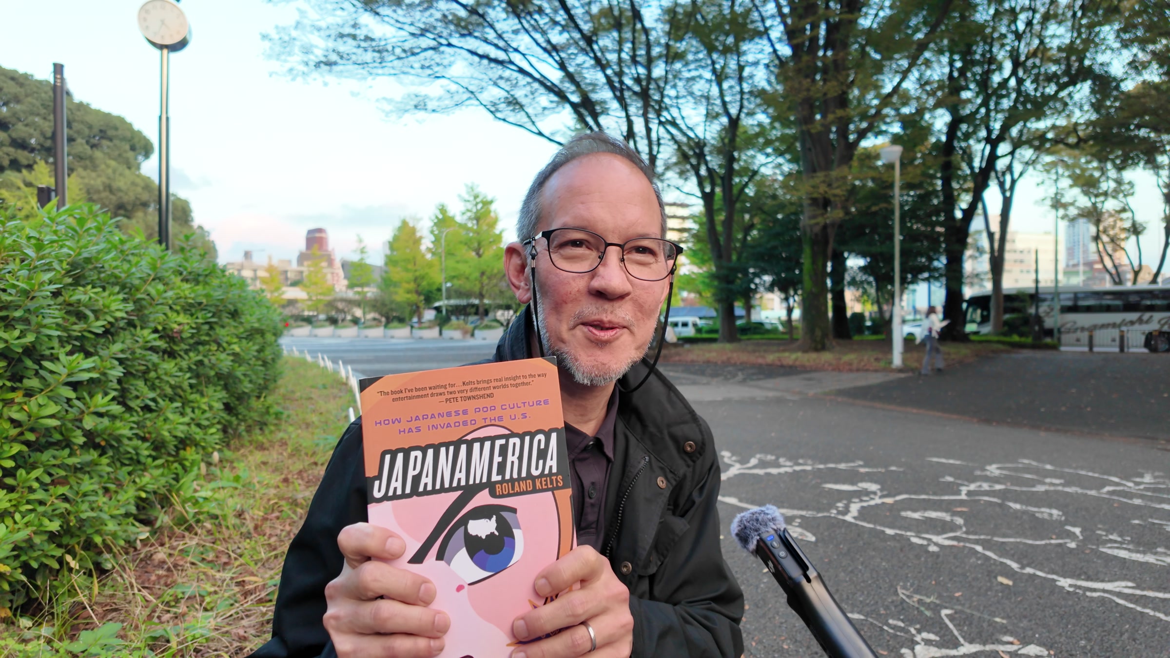 Roland Kelts smiling and holding up a copy of his book Japanamerica outdoors, with trees behind him