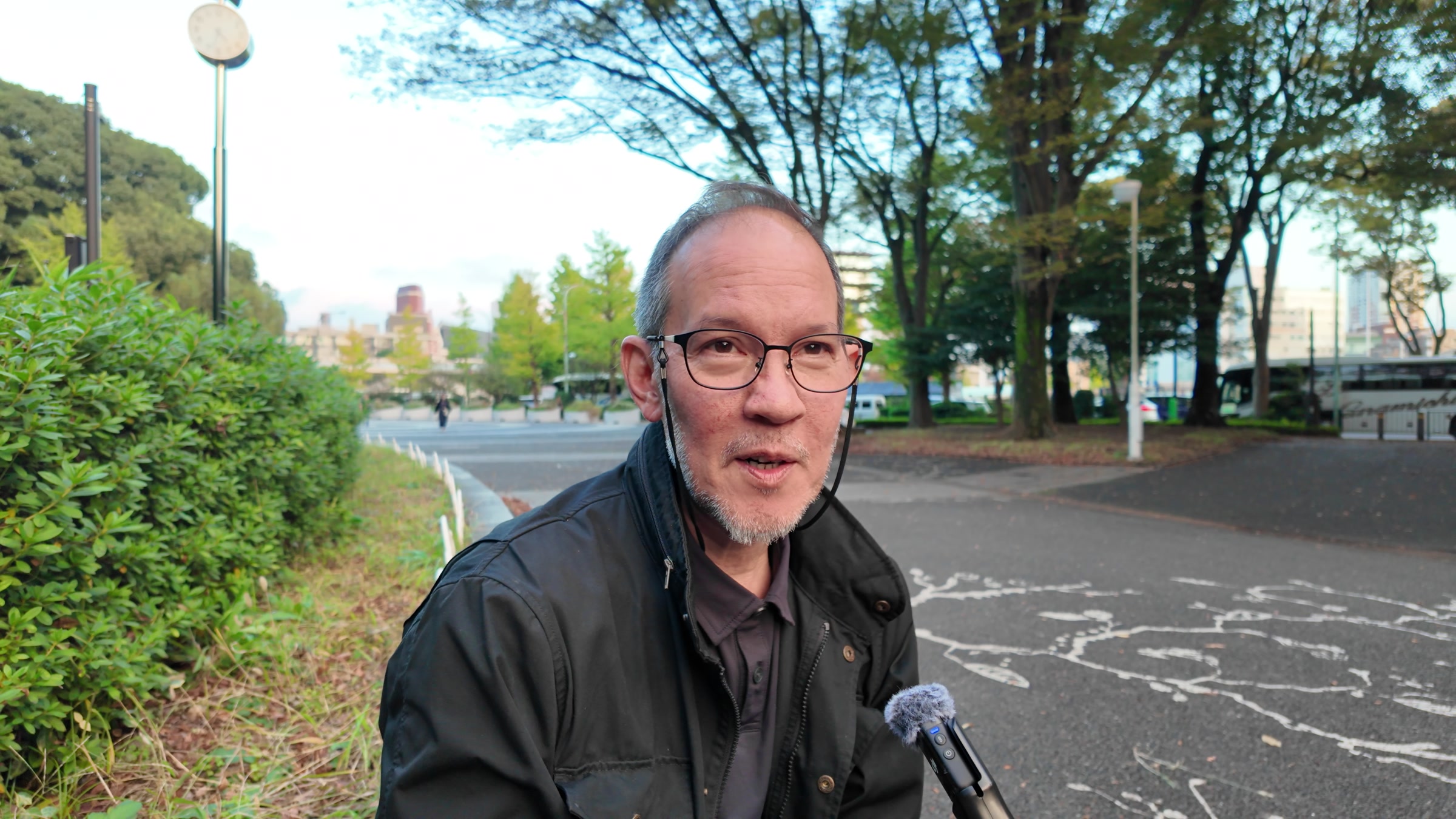 Roland Kelts speaking during an outdoor interview, with a hedge and a tree-lined road behind him