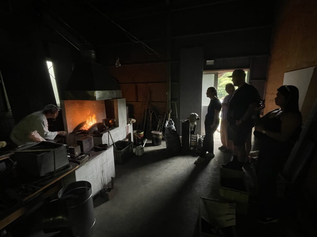 Visitors stand and watch as a Japanese swordsmith in white work clothes tends a charcoal forge in a dim workshop in Seki